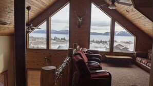 A cozy room with a sloped ceiling, large windows offering a view of a snowy landscape and mountains, a ceiling fan, and a fireplace. - Raspberry Bear Lake Lodge - 6 Bed 3 Bath. Great View Of The Lake! Perfect For Big Families! (Garden City)