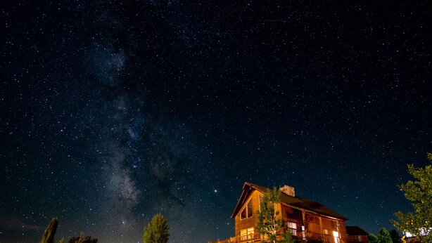 A large log cabin with a stone foundation, surrounded by trees and a clear night sky with visible stars and the Milky Way. - Raspberry Bear Lake Lodge - 6 Bed 3 Bath. Great View Of The Lake! Perfect For Big Families! (Garden City)