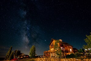 Una cabaña de troncos grande con cimientos de piedra, rodeada de árboles y un cielo nocturno despejado con estrellas visibles y la Vía Láctea.