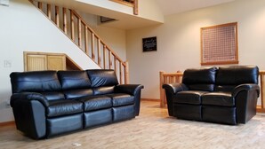 Un salon avec un canapé et un fauteuil en cuir noir, un escalier en bois et un store à carreaux.