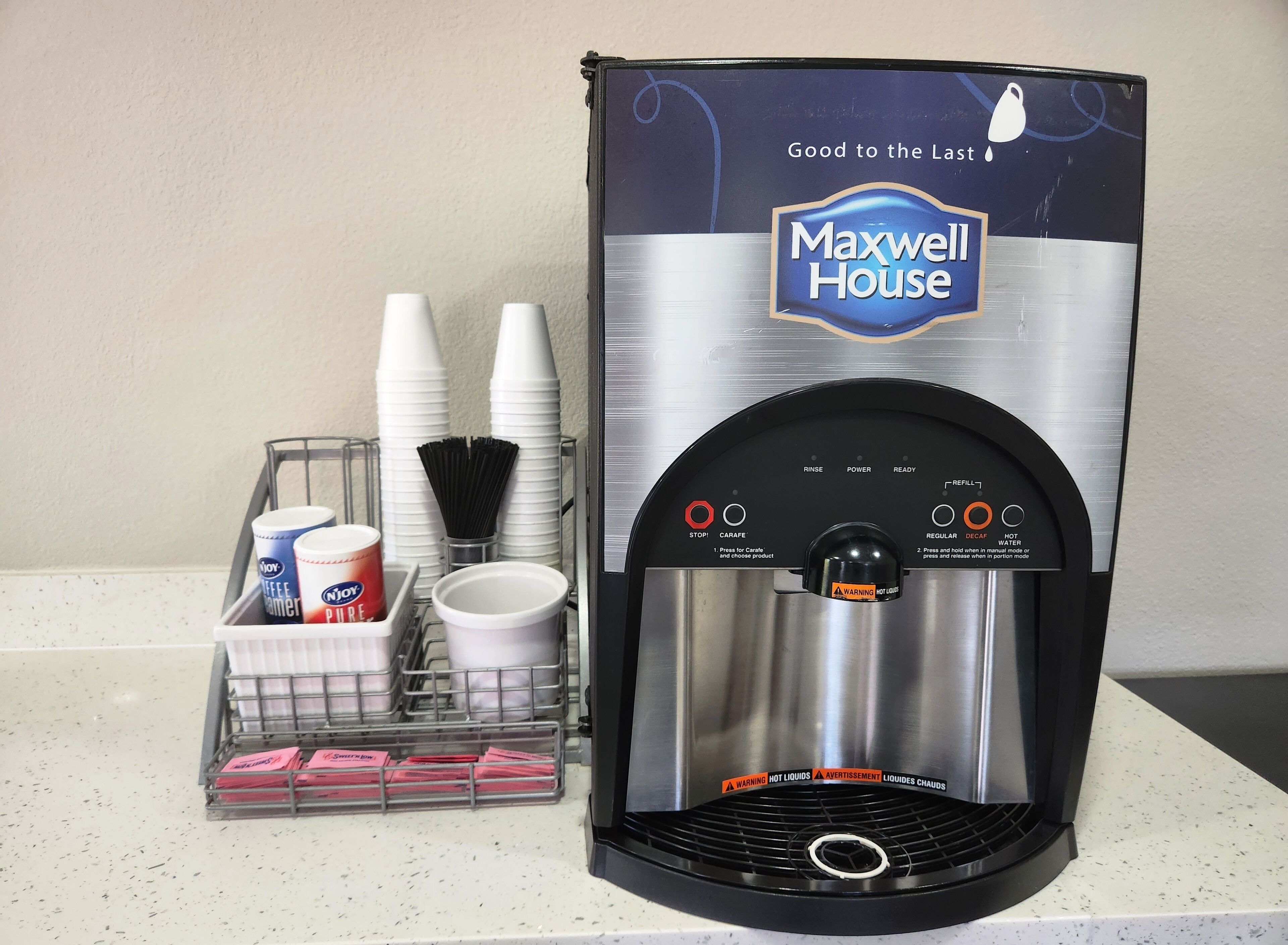 A water dispenser branded 'Maxwell House' with a stainless steel design, a cup holder, and a tray with disposable cups and a coffee cup.