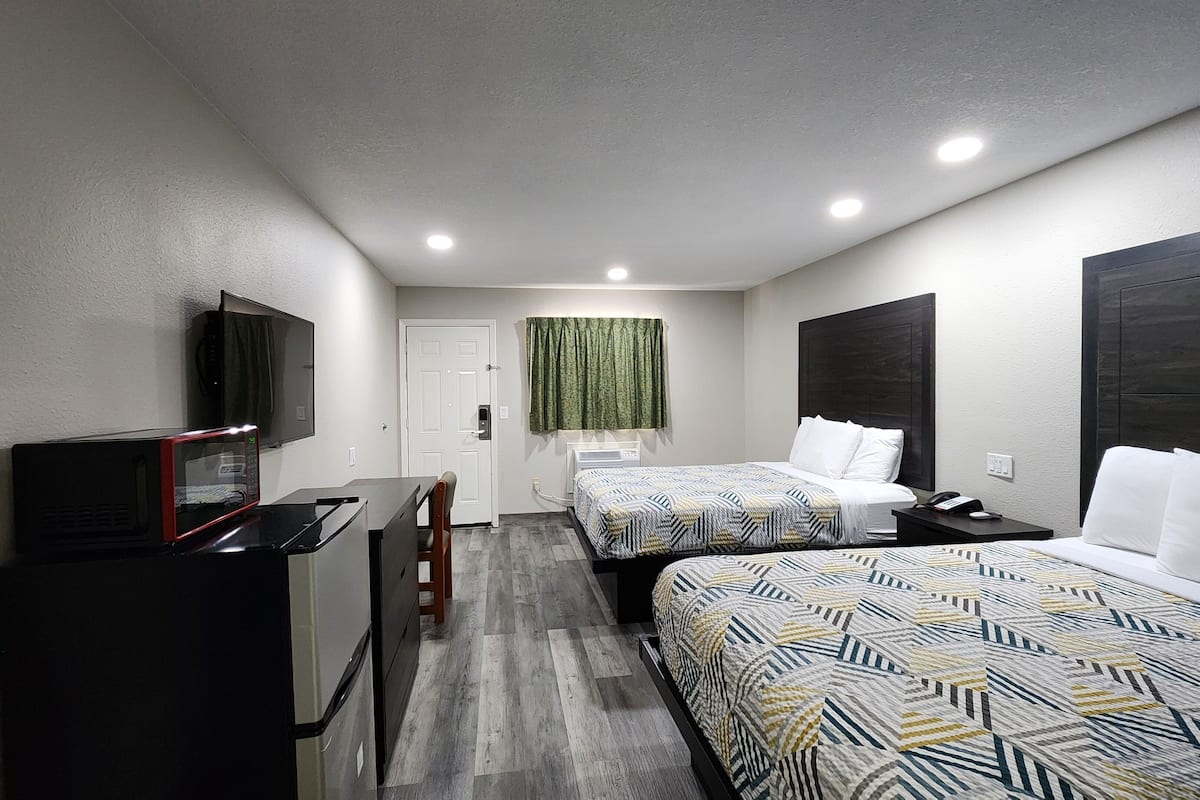 A hotel room with two beds, a desk with a microwave, a TV, and a window with a green curtain.