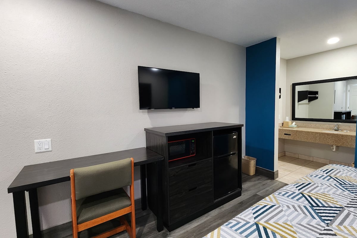 A hotel room with a desk, chair, TV, minibar, and a bed with a patterned bedspread.