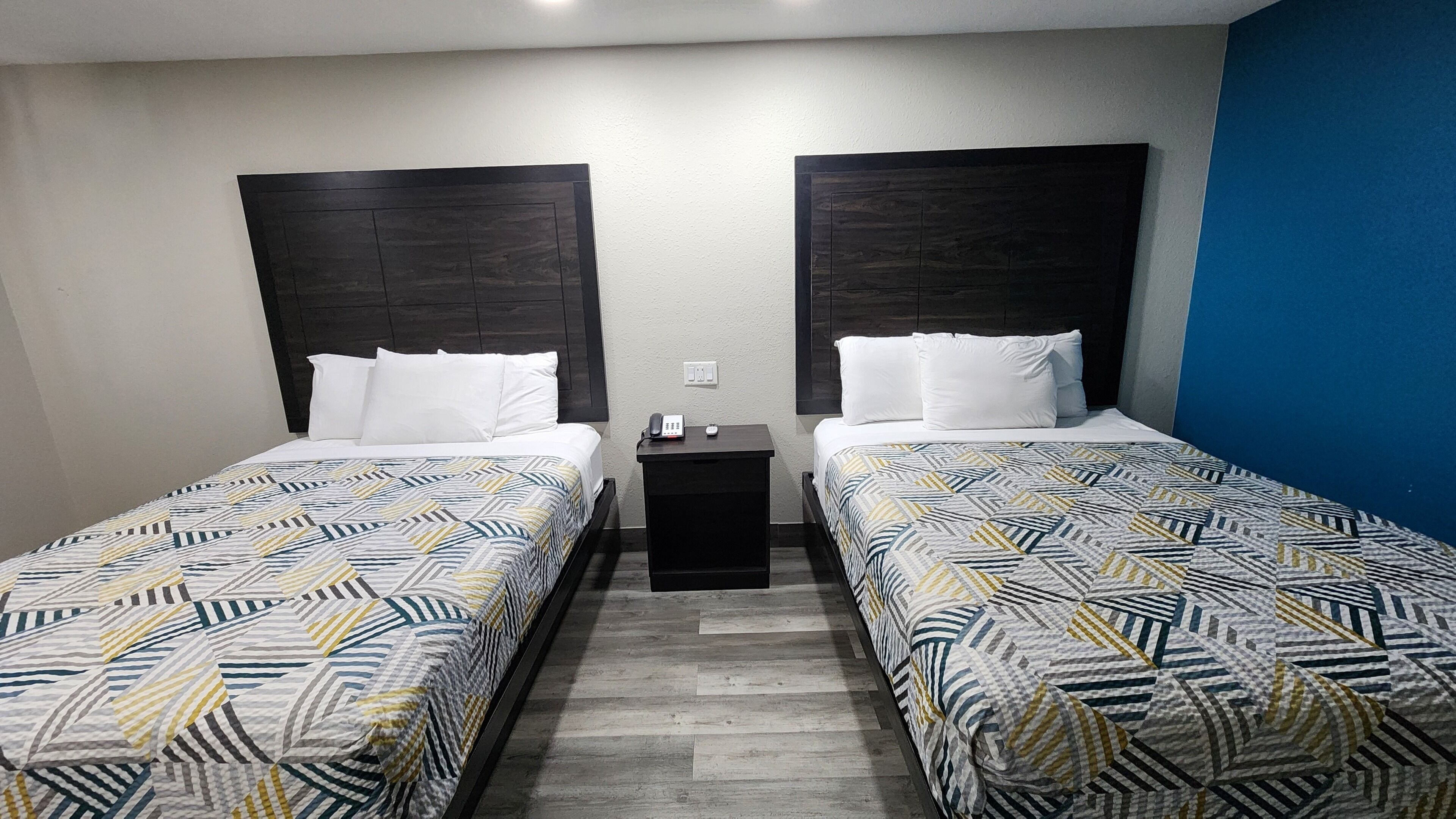 A hotel room with two beds, each with a patterned bedspread, a nightstand with a phone and a remote, and a wall switch.