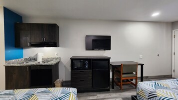 Una kitchenette compacta con microondas, un refrigerador pequeño y un lavamanos. Hay un televisor montado en la pared y una sola silla. La habitación tiene una distribución sencilla con dos camas.