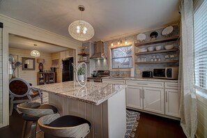A kitchen with a central island, bar stools, a chandelier, and a window with blinds. - Creekside Cottages Luxury King/The Nook (Blairsville)