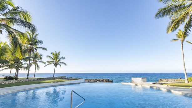 A swimming pool with a view of the ocean, surrounded by palm trees and a clear sky.