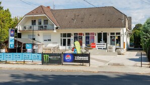 Ein zweigeschossiges Gebäude mit einem Ziegeldach und einem Balkon. Mehrere Schilder für Marken wie Pepsi, Balaton ABC und Monster Energy sind sichtbar.