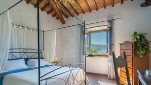 Uma cama com dossel e cortina, uma escrivaninha de madeira, uma cadeira, uma pequena mesa com abajur, e vista do exterior pela janela.
