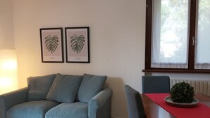 Una sala de estar con un sofá azul claro, una mesa de madera con mantel rojo, y dos láminas de plantas enmarcadas en la pared.