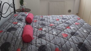 Una cama con colcha estampada y dos almohadas rojas, una mesita con una planta, y un radiador al fondo.