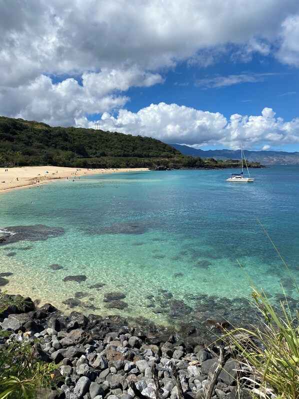 Une plage bordée d’une eau turquoise et limpide, un voilier et un premier plan rocheux.
