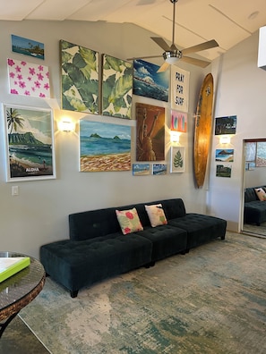 Un salon avec un canapé vert foncé, des œuvres d’art accrochées au mur, un ventilateur de plafond et une planche de surf.