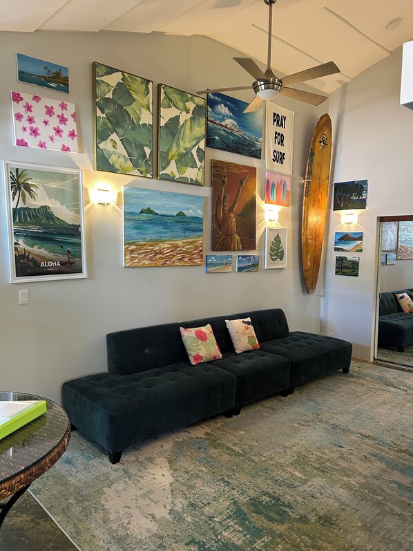 A living room with a dark green sofa, wall-mounted artwork, a ceiling fan, and a surfboard.