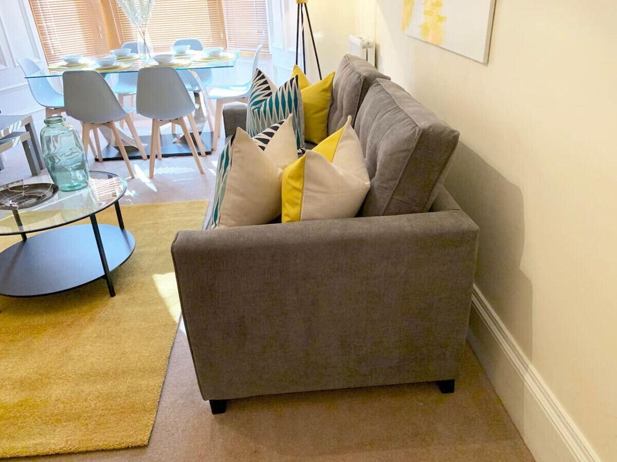 Ein Wohnzimmer mit grauem Sofa, gelb-grauen Kissen, einem Glastisch und einem gelben Teppich.