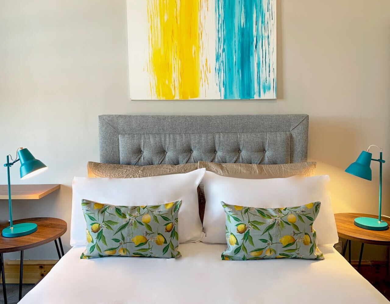 A hotel room with a tufted headboard, white bedding, and two matching pillows with a lemon pattern. There are two side tables with lamps, and a large abstract painting above the bed.