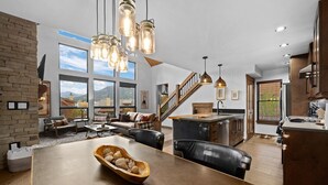 A spacious living area with a large wooden table, a stone fireplace, and a view of mountains through large windows. - Beautiful Views! Jacuzzi, Indoor/ Outdoor Fireplace, Steps to Lake Estes (Estes Park)