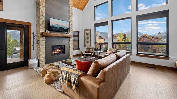 A spacious living room with a stone fireplace, large windows, and a high ceiling with exposed beams. - Beautiful Views! Jacuzzi, Indoor/ Outdoor Fireplace, Steps to Lake Estes (Estes Park)