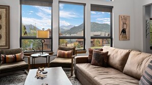 A spacious living room with large windows offering mountain views, a cozy seating area with a sofa and chairs, and a coffee table with decorative items. - Beautiful Views! Jacuzzi, Indoor/ Outdoor Fireplace, Steps to Lake Estes (Estes Park)