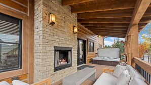 A covered outdoor seating area with a stone fireplace, wooden deck, and a hot tub. - Beautiful Views! Jacuzzi, Indoor/ Outdoor Fireplace, Steps to Lake Estes (Estes Park)