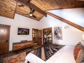 A spacious bedroom with a large bed, wooden ceiling beams, a ceiling fan, and a nightstand with wine glasses. - Relax in Luxury - Jacuzzi, Indoor/Outdoor Fireplace (Estes Park)