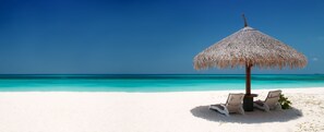 A beach with a thatched umbrella and two lounge chairs.