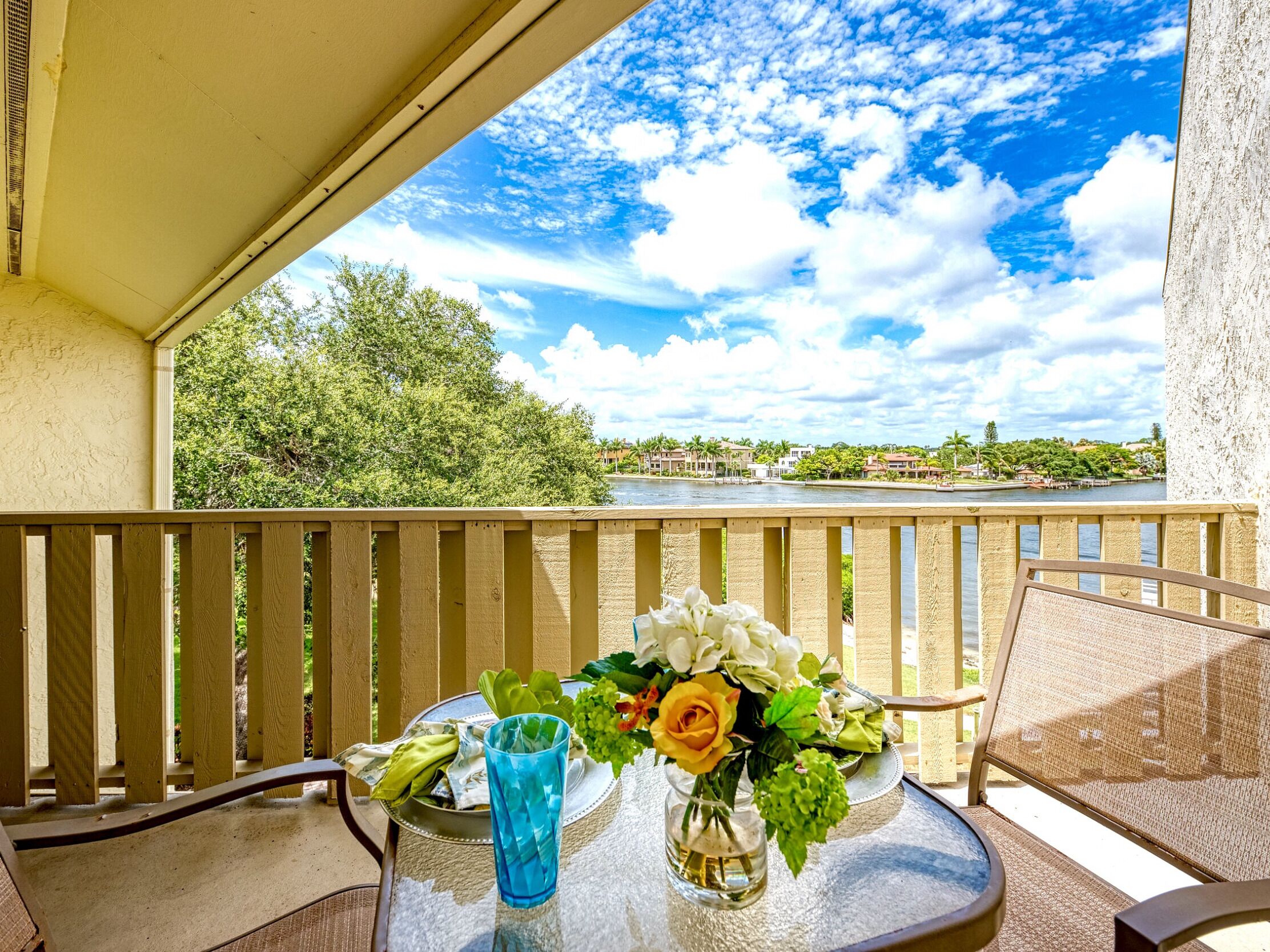 A balcony with a table set for two, overlooking a river with a view of houses and greenery.