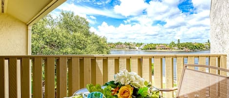 A balcony with a table set for two, overlooking a river with a view of houses and greenery.