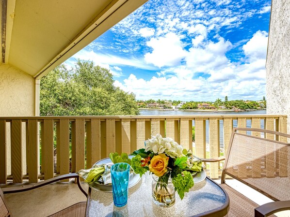 A balcony with a table set for two, overlooking a river with a view of houses and greenery.