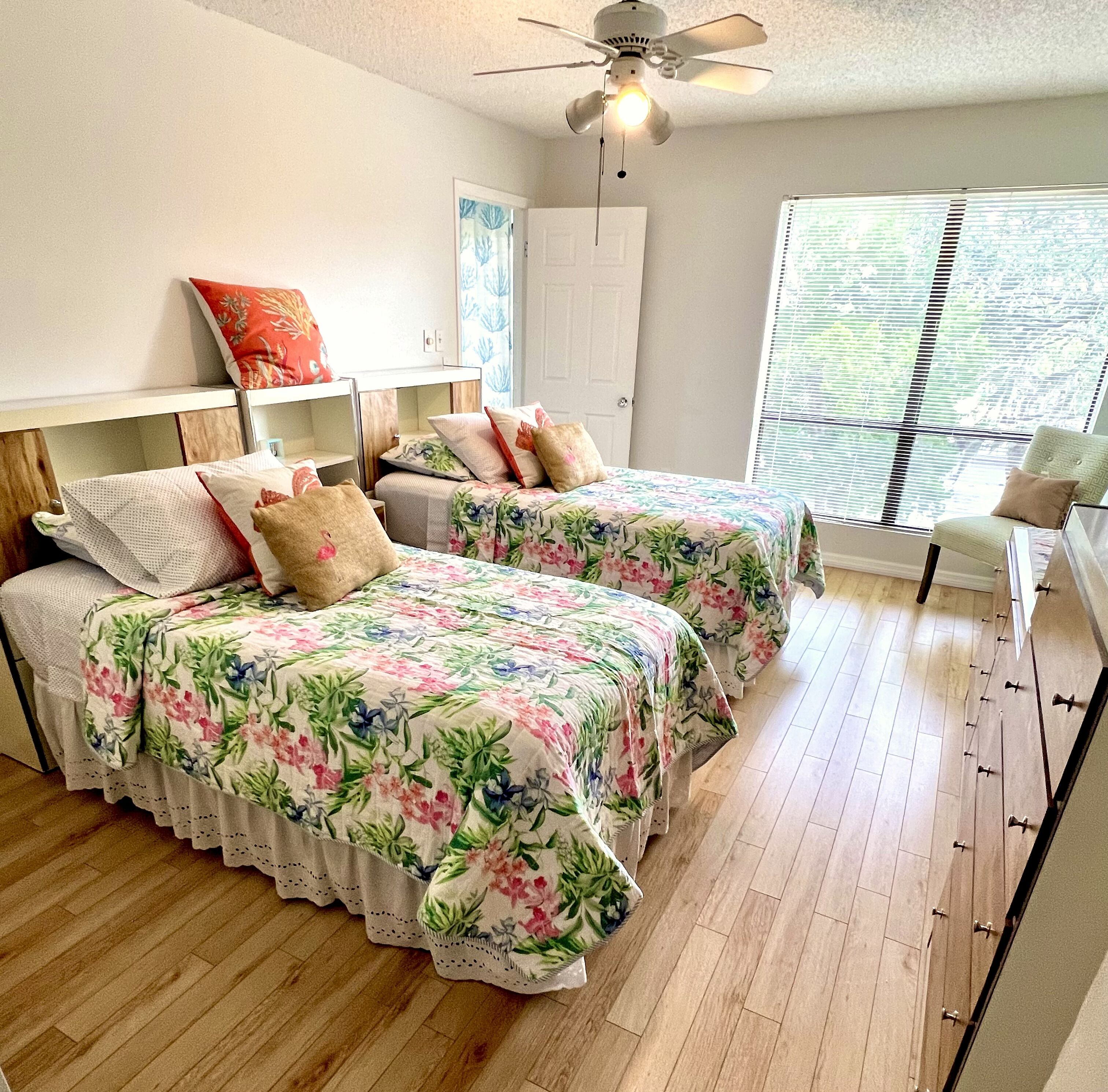 A bedroom with two beds, wooden flooring, a ceiling fan, and a window with blinds.