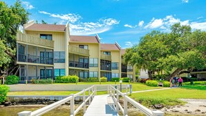 A wooden bridge leading to a building with multiple balconies and a grassy area with trees. - Luxury Waterfront Views / Pool / 1400sf / Minutes to Beach (Siesta Key)
