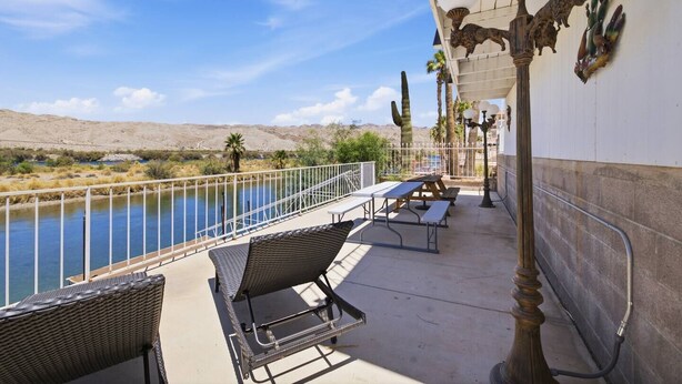 A balcony with wicker chairs, a table, and a railing overlooking a body of water and desert landscape.
