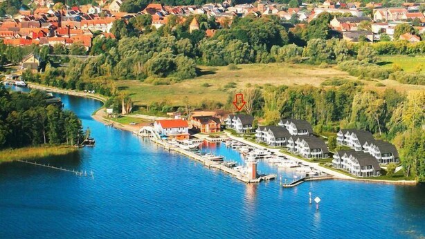An aerial view of a lakeside marina with docked boats, a small pier, and residential buildings in the background.