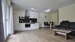 Eine moderne Küche mit weißen Schränken, einer Mikrowelle und einem Herd. Es gibt einen Essbereich mit Tisch und Stühlen. Im Wohnbereich befinden sich eine schwarze Couch und ein Couchtisch aus Holz.