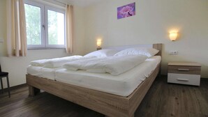 Ein ordentlich bezogenes Bett in einem Zimmer mit Holzboden, einem Fenster mit Vorhängen, einem Nachttisch mit Lampe und einer Wandleuchte.