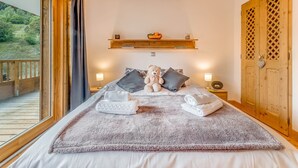 A bedroom with a large bed, two bedside lamps, a wooden shelf with items, and a view of a balcony. - 4* Apartment at Slopeside, Sleeps 8, Wi-Fi Included (Champagny-en-Vanoise)