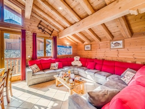 Ein gemütliches Blockhaus-Interieur mit einem großen Ecksofa, einem Holzespeisetisch mit Frühstücksgebäck und Blick ins Freie durch eine Glastür.