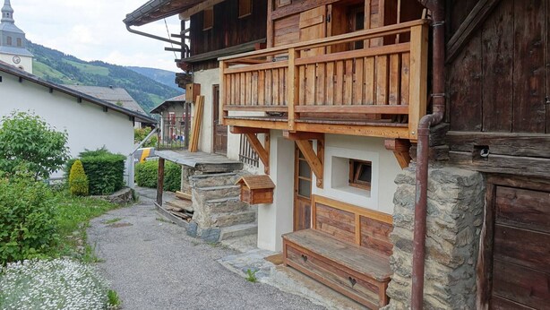 A traditional wooden house with a stone foundation, a wooden balcony, and a gravel driveway.