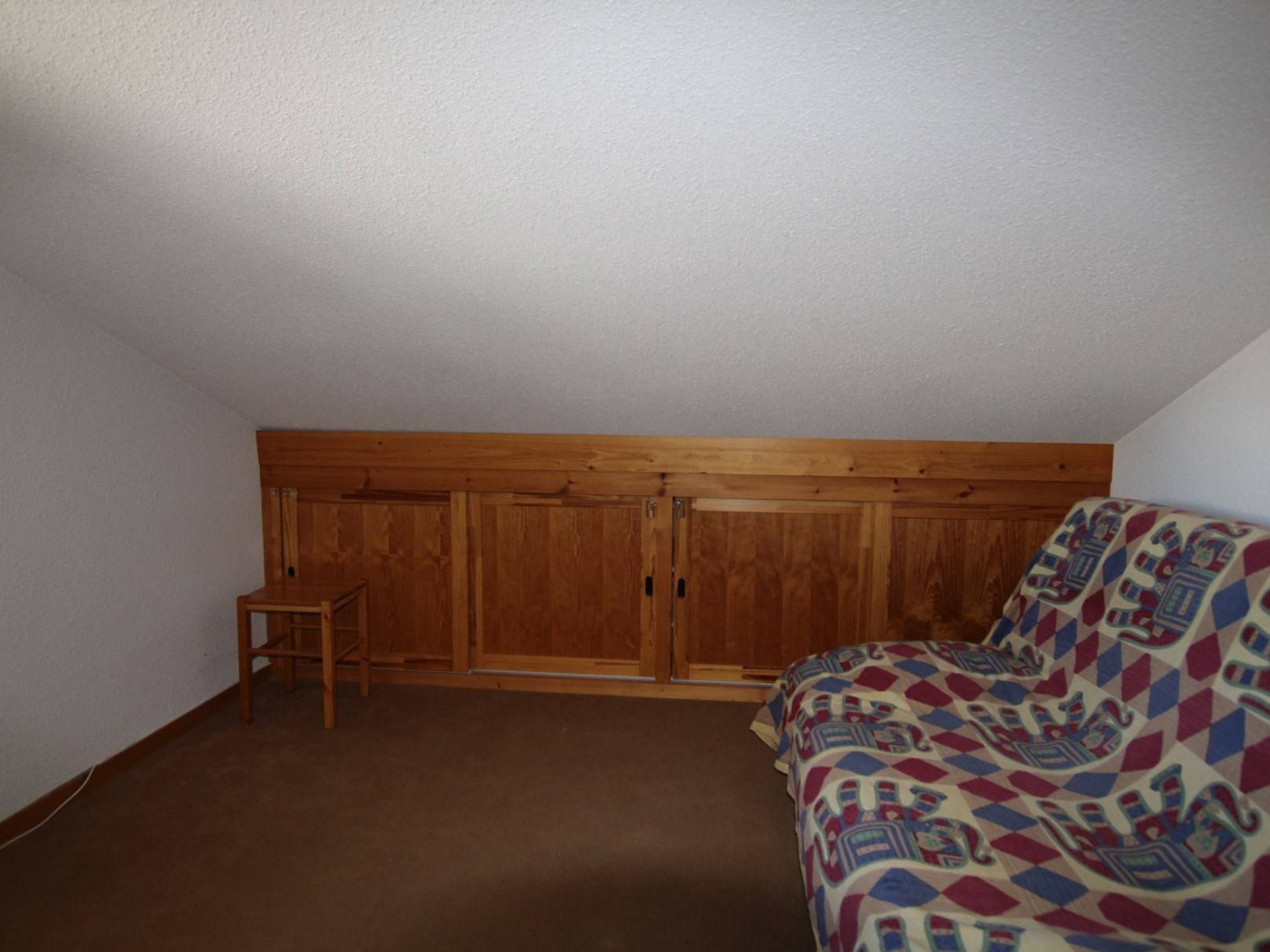 Una stanza piccola e poco illuminata, con soffitto spiovente, un armadio in legno, una sedia di legno e una coperta con motivi decorativi.