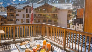Ein Balkon mit Frühstücksangeboten, darunter Früchte, Gebäck und Getränke, mit Blick auf einen Bergort mit Holzbauten und einer Flagge.