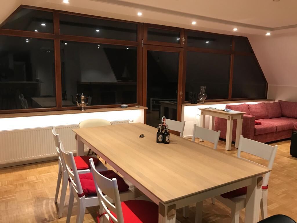 Un coin repas avec une table en bois, des chaises blanches et un coussin rouge. On peut voir deux bouteilles sur la table, et un canapé rose est visible à l’arrière-plan.