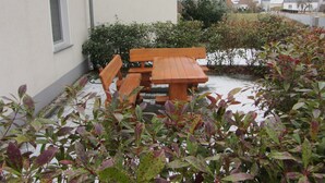 Ein Holzbänkchen und Tisch vor einem Gebäude mit einem Fenster und einem Hecken im Vordergrund.