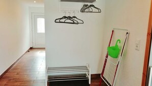 Ein Flur mit einer weißen Tür, einem Kleiderständer mit schwarzen Kleiderbügeln, einem grünen Korb und einem rot-weißen Klappstuhl.