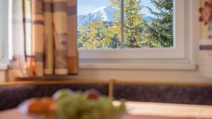 A dining table with a bowl of fruit, a window with a view of a mountain, and books on the side. - Comfortable Apartment “Treyer 3 Typ B” with Mountain View, Balcony & Garden (Terenten / Pustertal)