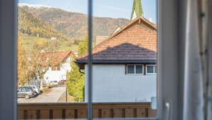 On aperçoit, à travers une fenêtre, une maison avec un toit en pente, une cheminée bien visible et, au loin, le clocher d’une église.