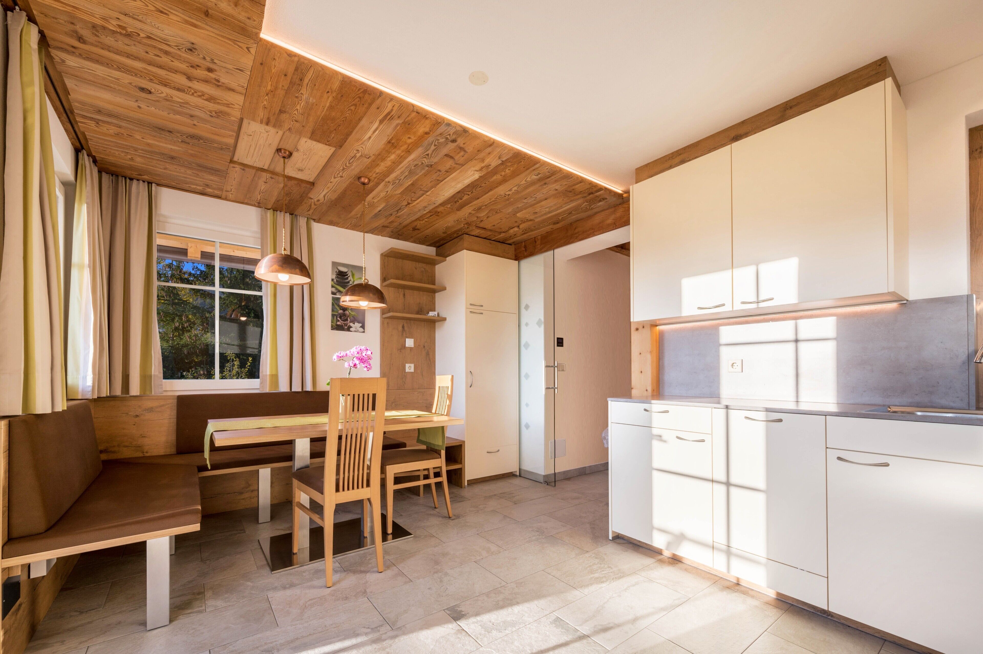A modern kitchen with wooden beams, a dining area with a bench, and a view of greenery outside.