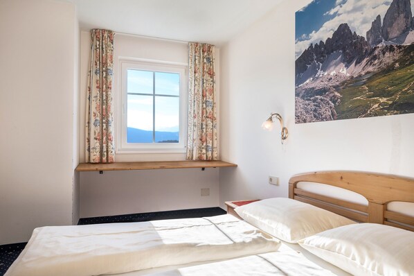 A bedroom with a wooden headboard, a window with floral curtains, a bed with white bedding, and a mountain landscape painting on the wall.