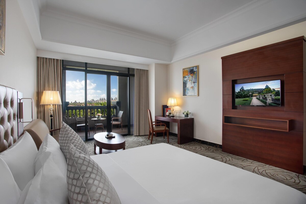 Habitación de hotel con cama, escritorio, silla, televisor y balcón con vistas.