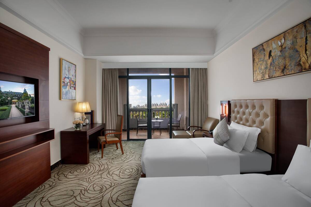 A hotel room with two beds, a desk, a TV, and a balcony with a view.