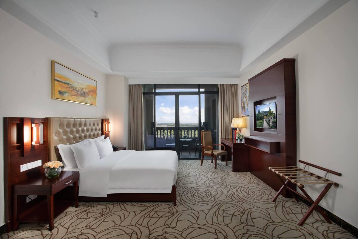 A hotel room with a large bed, a desk with a lamp, a TV, and a balcony with a view.